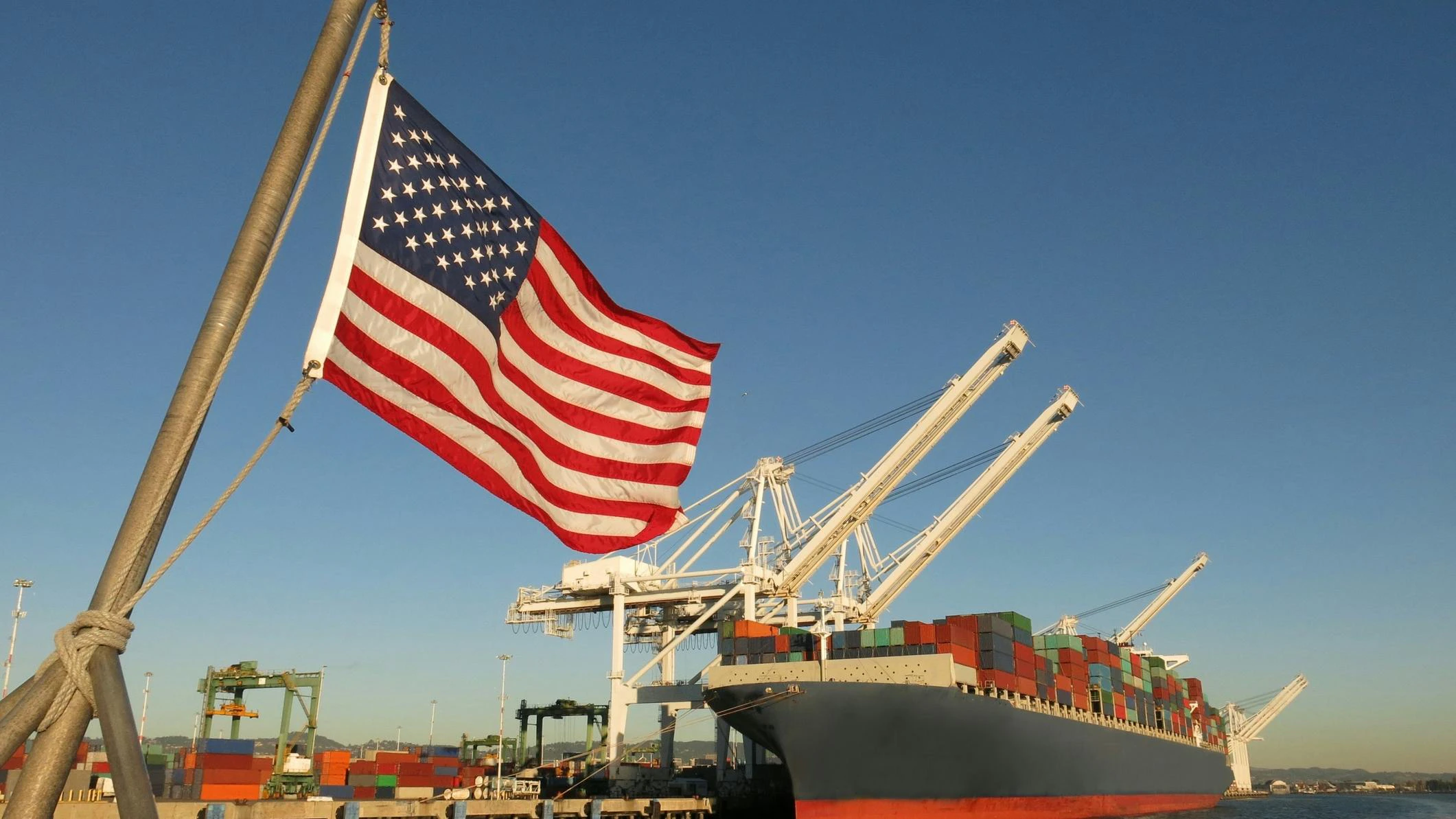 An American flag waves at a US port, where a cargo ship is docked beneath a crane on a blue sky day.