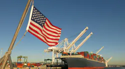 An American flag waves at a US port, where a cargo ship is docked beneath a crane on a blue sky day. An American flag waves at a US port, where a cargo ship is docked beneath a crane on a blue sky day.