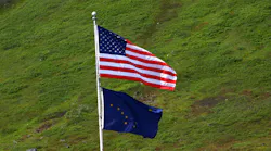 US and Alaska flag flying on flag pole with grassy hill in background.