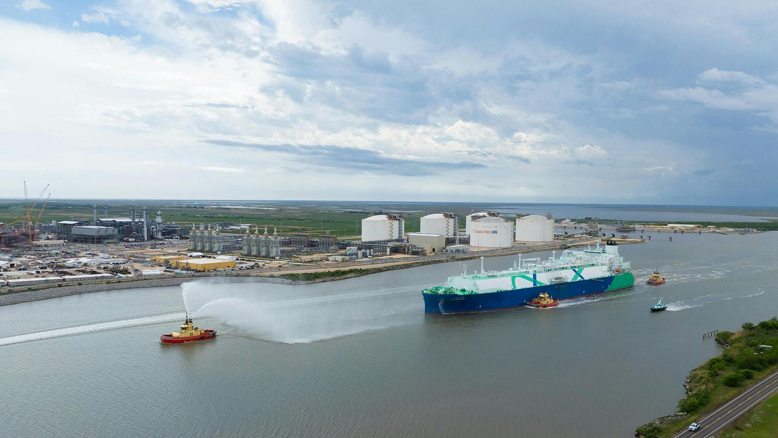 Tanker containing first shipment from the Golden Pass LNG project is guided by tug vessels through a canal waterway out to open sea.