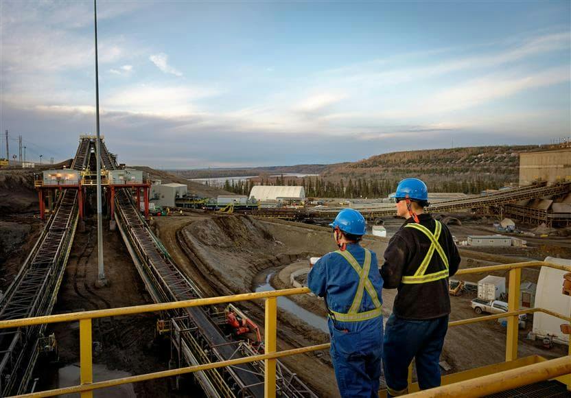 Two individuals in hardhats and blue coveralls on a bridge overlooking equipment at a Canadian oil sands production site with a lake and forest in the background.