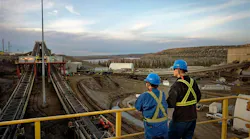 Two individuals in hardhats and blue coveralls on a bridge overlooking equipment at a Canadian oil sands production site with a lake and forest in the background.