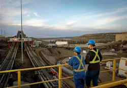Two individuals in hardhats and blue coveralls on a bridge overlooking equipment at a Canadian oil sands production site with a lake and forest in the background.