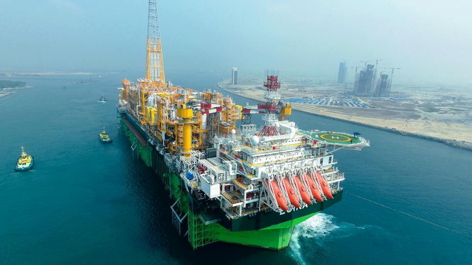 Large floating oil and gas production, storage and offloading vessel shown near the coastline escorted by smaller vessels